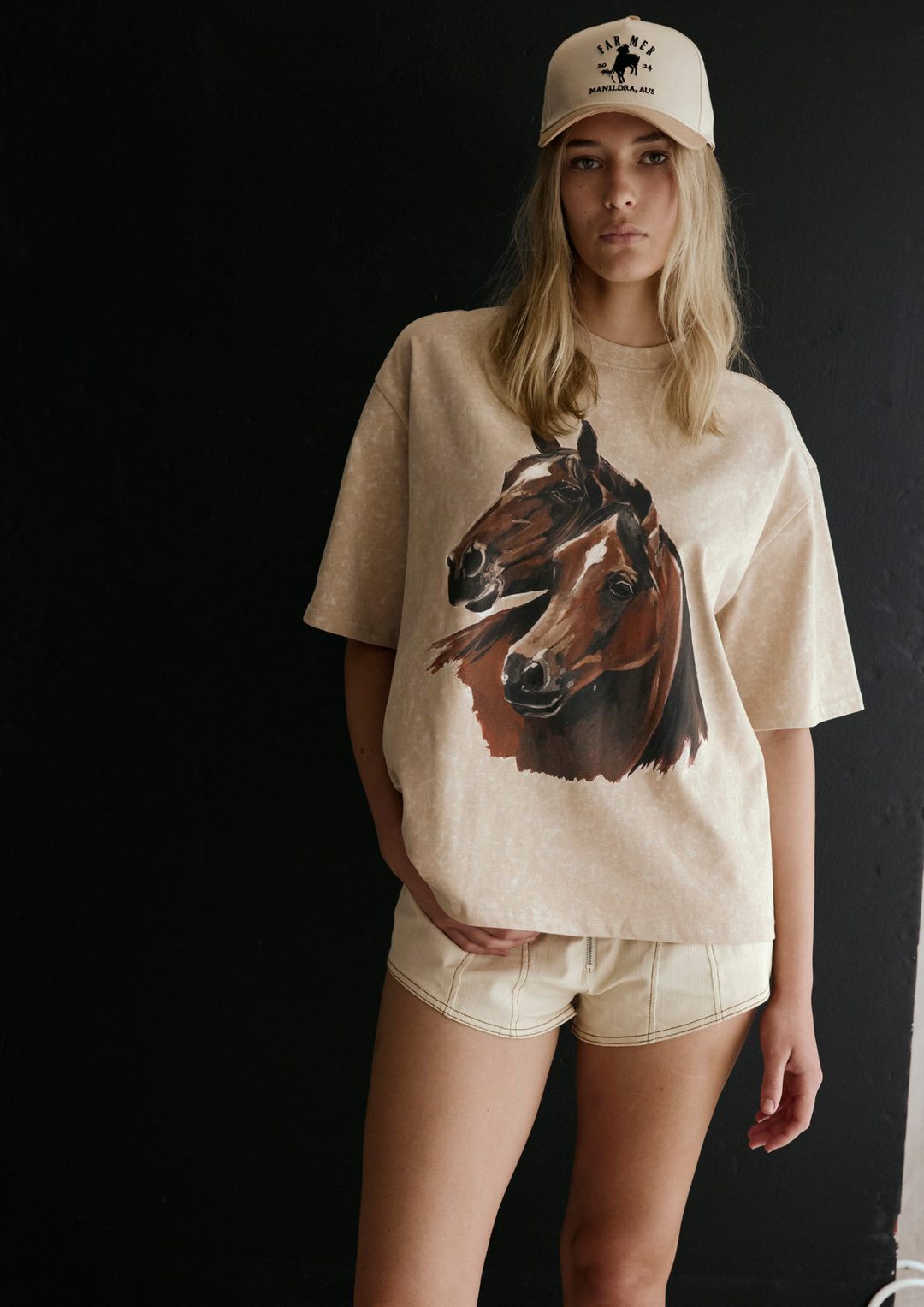 Person wearing a beige t-shirt with a horse graphic and a cap on a black background
