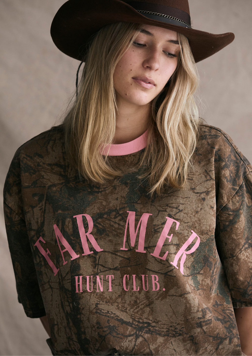 Person wearing a camouflage shirt with pink text and a brown hat on a neutral background