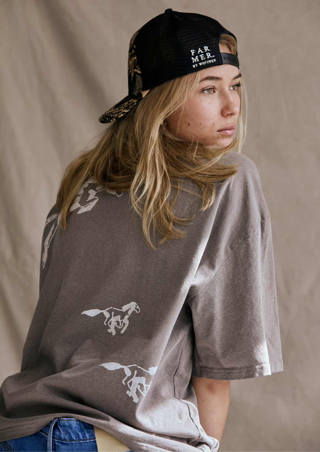 Person wearing a grey t-shirt with horse designs and a black cap on a beige background