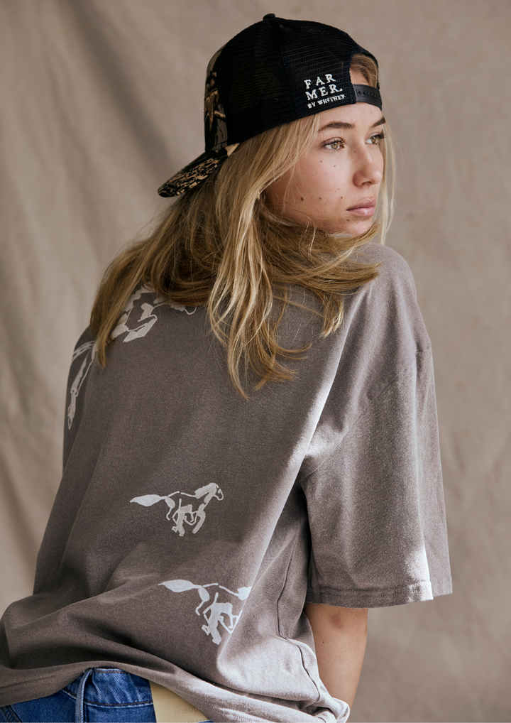 Person wearing a grey t-shirt with horse designs and a black cap on a beige background