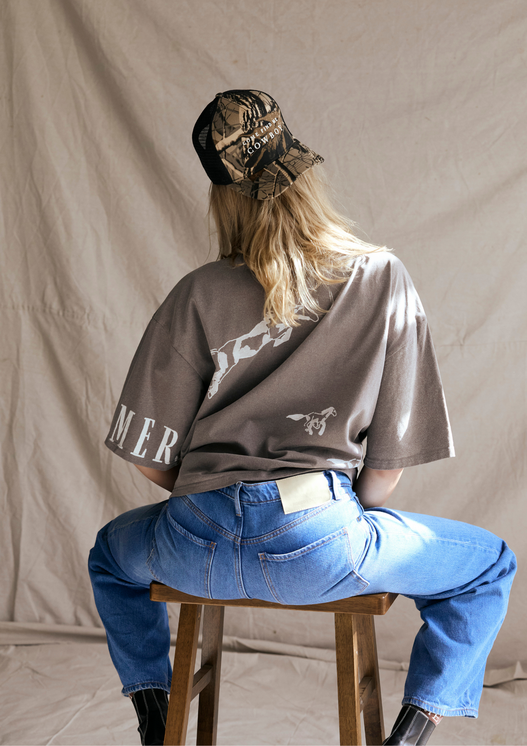 Person wearing a grey t-shirt with text and graphics, blue jeans, and a camouflage cap, sitting on a wooden stool.
