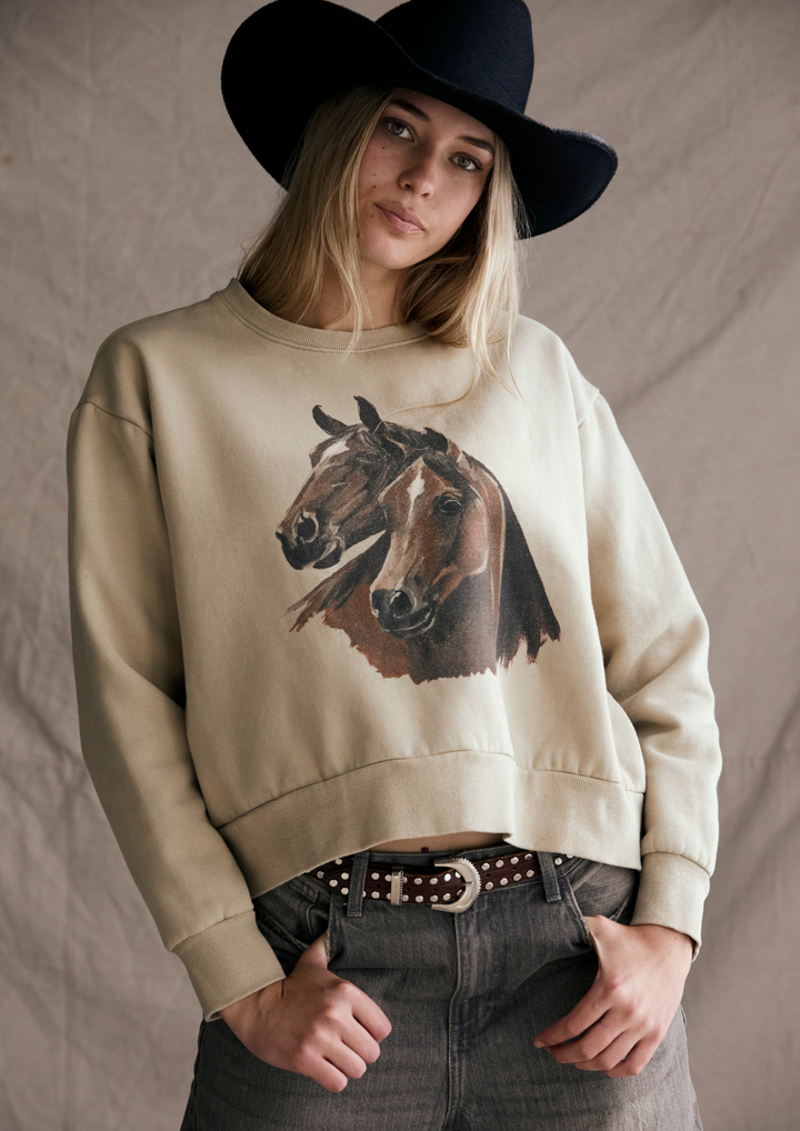 Person wearing a beige sweatshirt with a horse graphic, black hat, and dark jeans against a neutral background