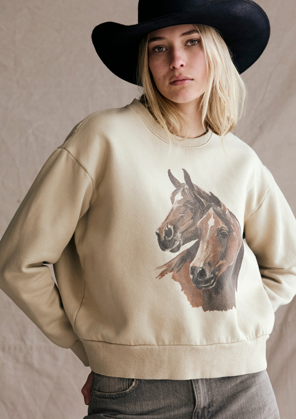 Person wearing a beige sweatshirt with a horse graphic and a black hat.